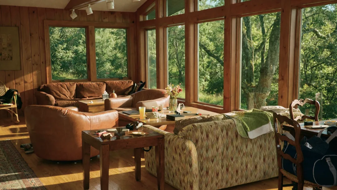 Sunlit living room with leather couches, floor-to-ceiling windows facing the woods